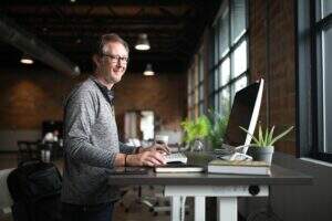Negócio sustentável: imagem de ambiente industrial que parece ser um escritório de trabalho. Foco da imagem: homem de pele clara e cabelo grisalho está em pé, em frente à mesa de trabalho com computador, celular, bloco de anotações, copo de café e algumas plantas verdes.