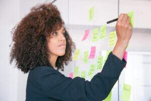 Mulher negra de cabelos cacheados e castanhos, anotando em post-its coloridos. Ela veste uma camisa azul marinho e simula planejar campanhas que vendem o ano todo.