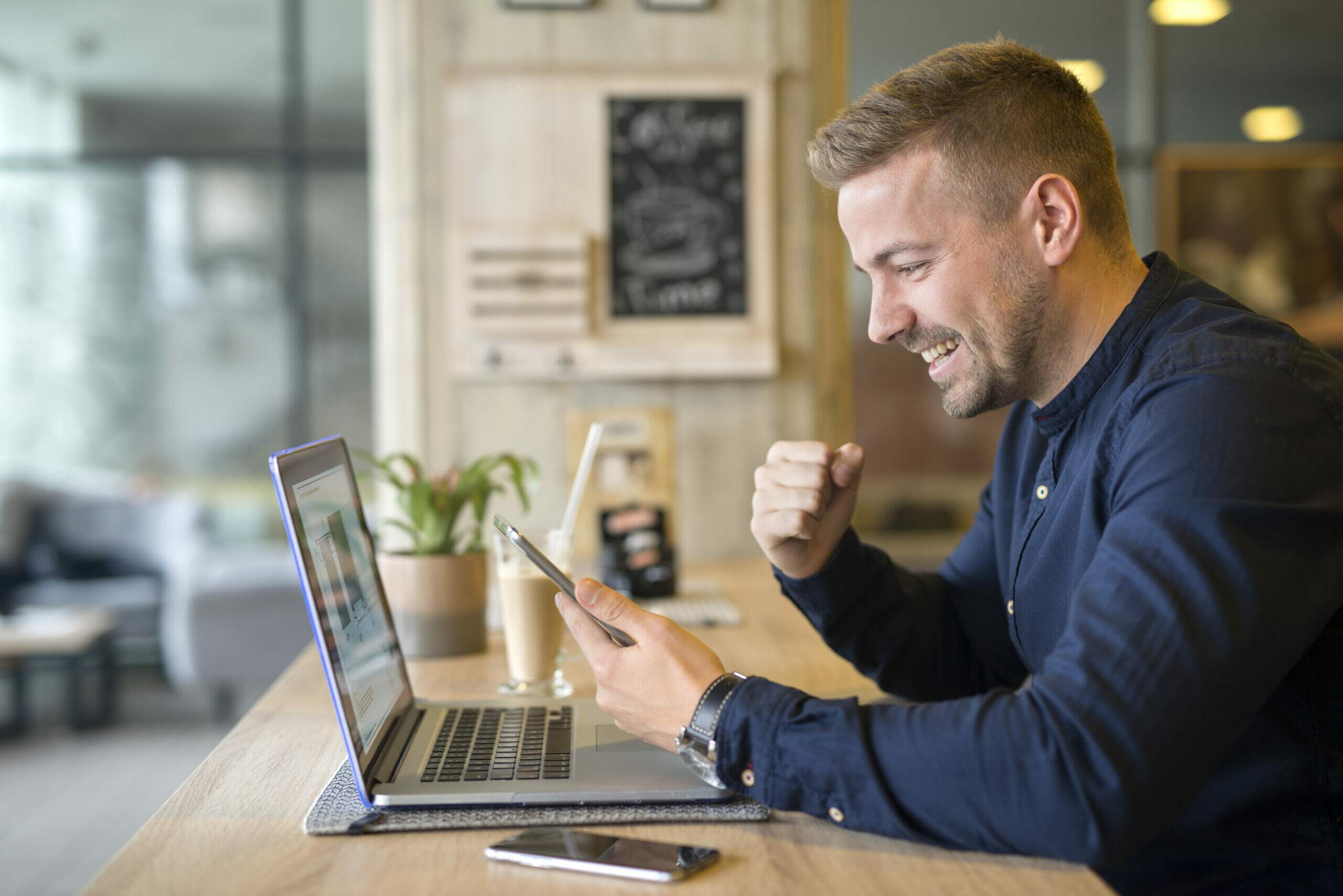 Catálogo digital vendizap - Homem branco com cabelo loiro, veste camisa social e comemora resultados que aparecem no computador e celular.